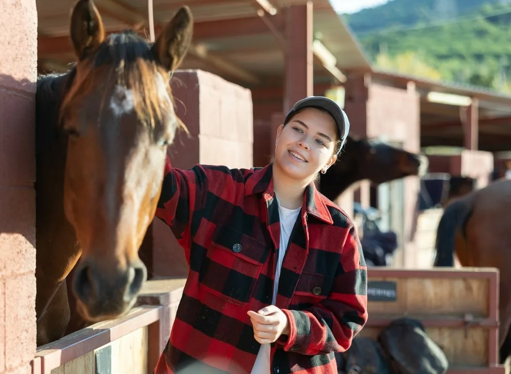 Caring young girl in plaid shirt cleaning and petting purebreed horse with in a countryside club.