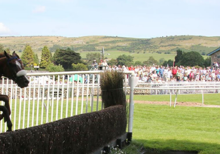 Jockey on horseback leaping over a hurdle at Cartmel Racecourse with spectators and rolling hills