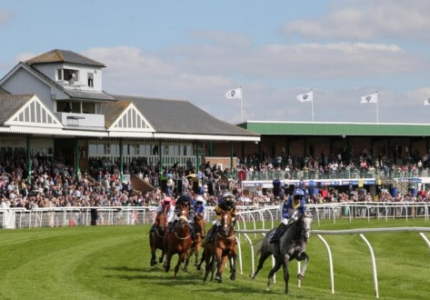 Races at Catterick with jockeys on the track and spectators gathered in the grandstand under a clear sky